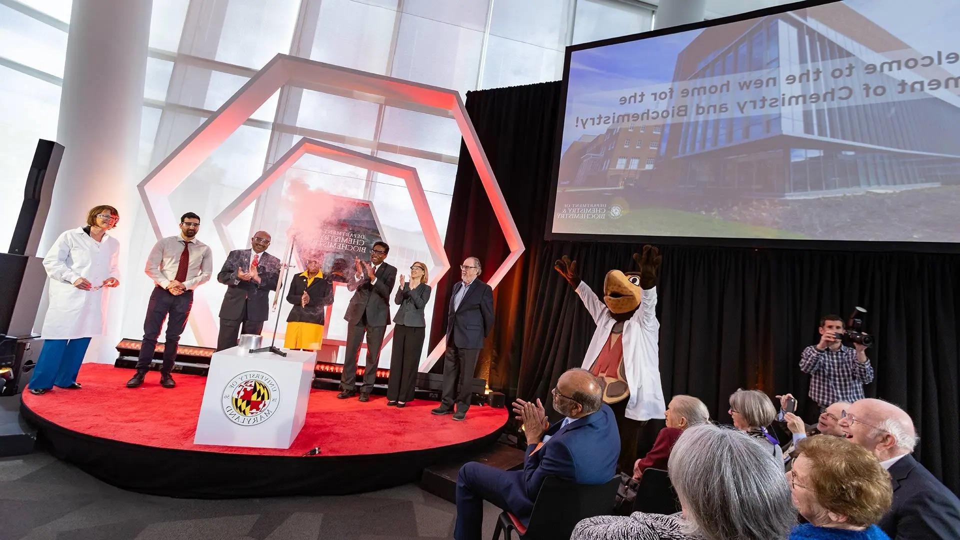 A chemical reaction illuminates a tiny representation of Testudo as the life-size UMD mascot and other participants at a dedication ceremony for the new Chemistry Building applaud. From left: Professor Emeritus Phil DeShong; Senior Vice President and Provost Jennifer King Rice; President Darryll J. Pines; Maryland House of Delegates Speaker Adrienne A. Jones; College of Computer, Mathematical, and Natural Sciences Dean Amitabh Varshney; doctoral student Hector Cein Mandujano and Department of Chemistry